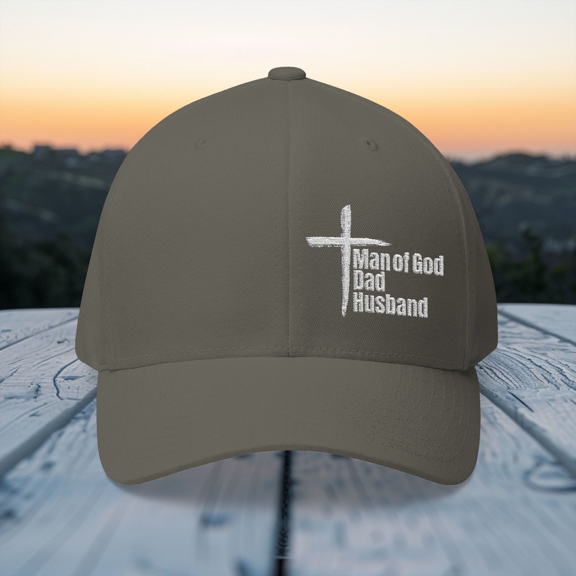 Man of God Dad Husband Embroidered Flexfit Hat displayed outdoors with a sunset backdrop.