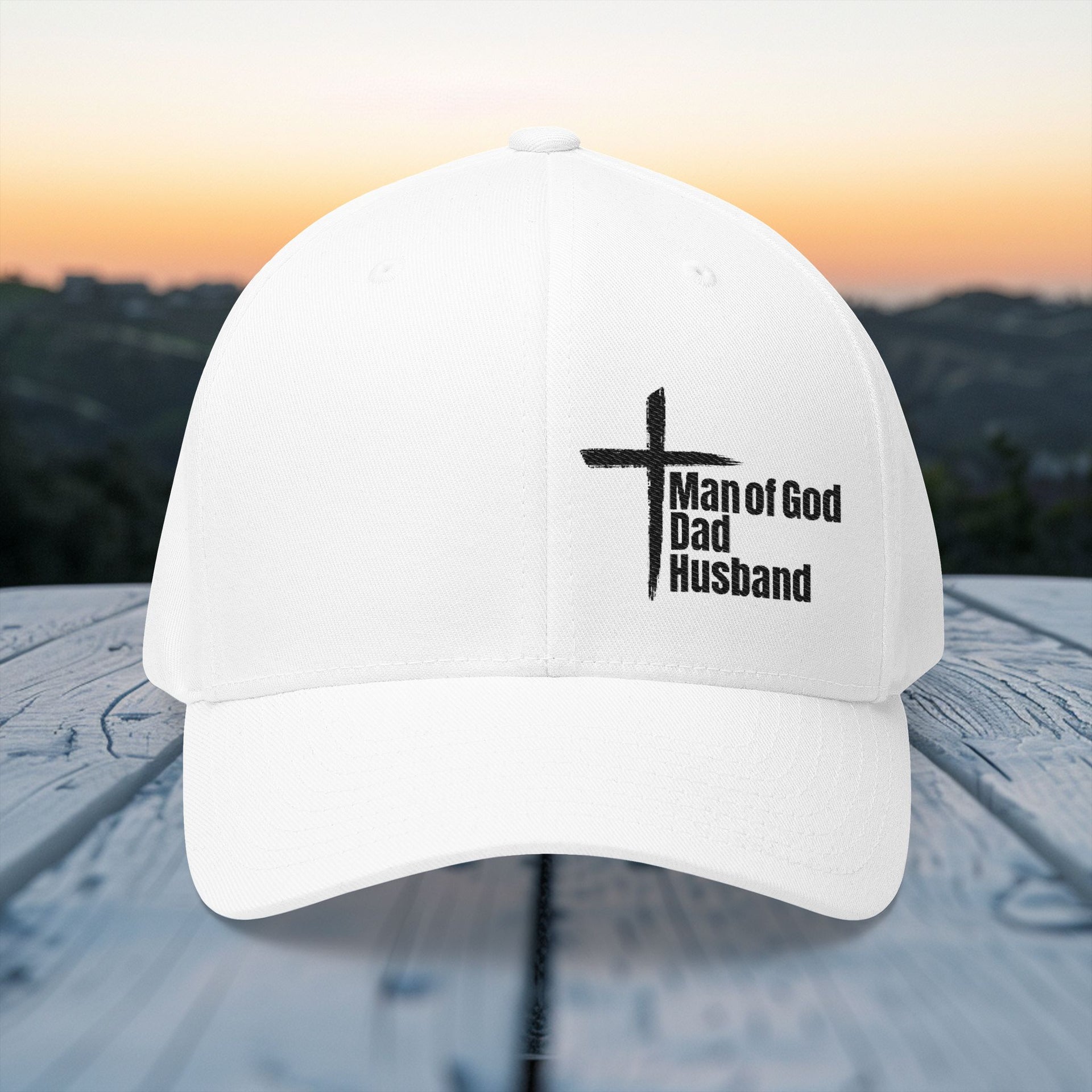 Man of God Dad Husband Embroidered Flexfit Hat in white displayed on a wooden table with a scenic background.