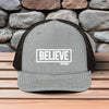 Believe In Christ Snapback Hat on wooden table with stone background showcases bold faith statement.