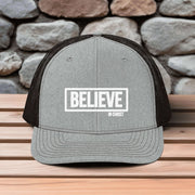 Believe In Christ Snapback Hat on wooden table with stone background showcases bold faith statement.