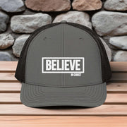 Believe In Christ Snapback Hat featuring embroidered design on a gray trucker hat against a stone background.