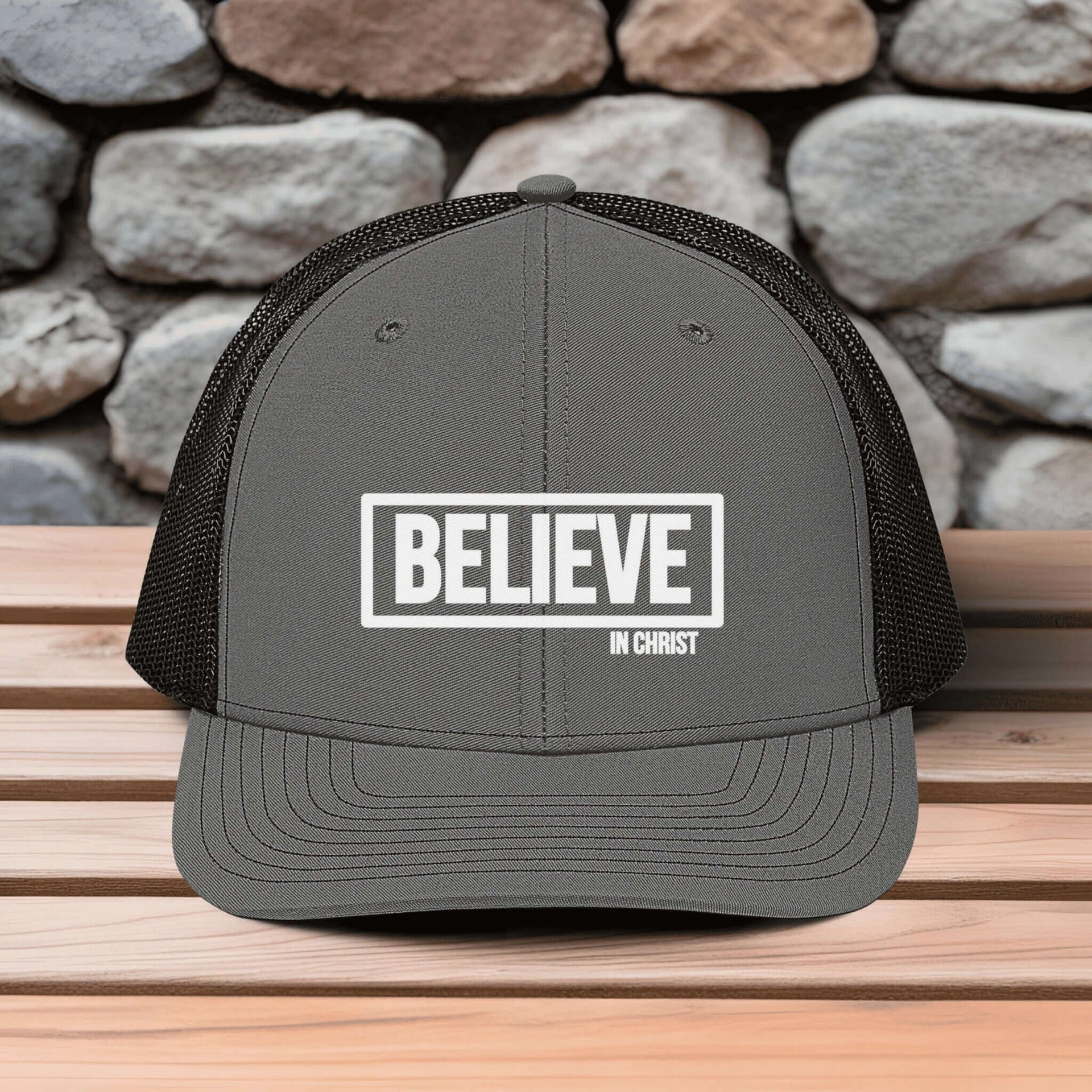 Believe In Christ Snapback Hat featuring embroidered design on a gray trucker hat against a stone background.