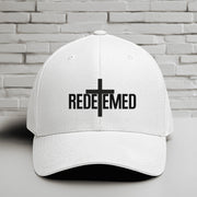 Redeemed Embroidered Flexfit Hat with a white base and black lettering showcasing the word 'REDEEMED' and a cross design.