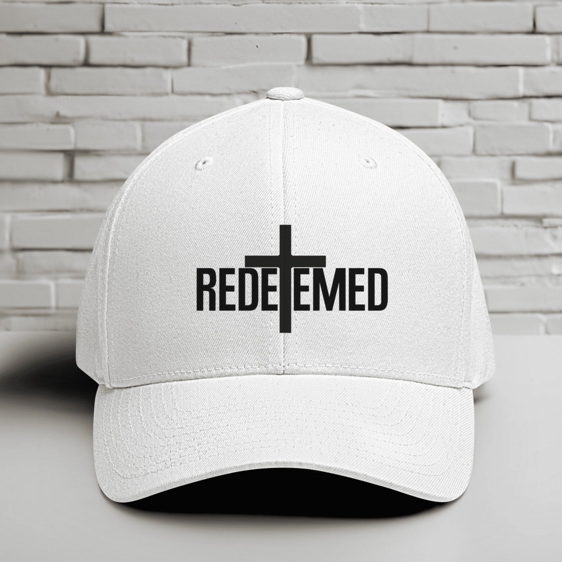 Redeemed Embroidered Flexfit Hat with a white base and black lettering showcasing the word 'REDEEMED' and a cross design.