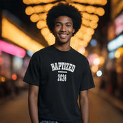 A young man smiles while wearing a black Baptized 2026 T-Shirt in a vibrant, illuminated street setting.