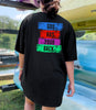 Girl wearing a God Has Your Back T-shirt with colorful block text on the back, standing near kayaks.