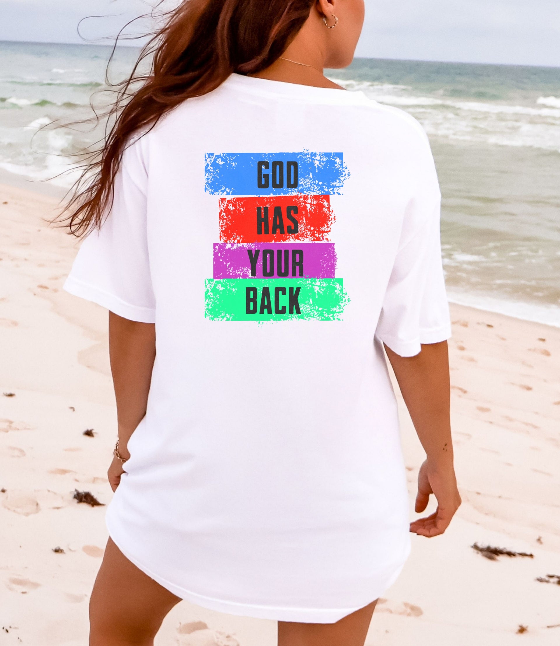 God Has Your Back T-shirt featuring colorful text design on the back, worn on the beach.