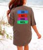 Back view of a woman wearing a God Has Your Back T-shirt on the beach with colorful graphic design.