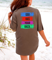 Back view of a woman wearing a God Has Your Back T-shirt on the beach with colorful graphic design.