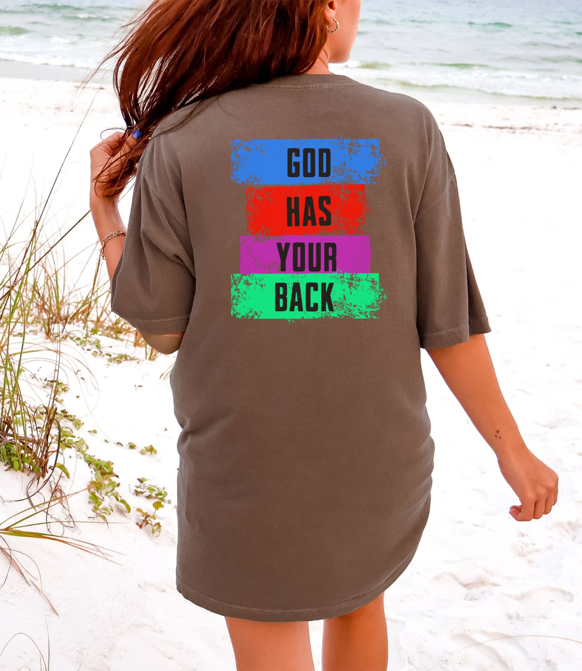 Back view of a woman wearing a God Has Your Back T-shirt on the beach with colorful graphic design.