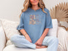 She Is Mom Floral Typography T Shirt worn by a woman sitting on a couch with soft fabric and a stylish design.