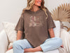 She Is Mom Floral Typography T Shirt worn by a woman sitting comfortably, showcasing a stylish floral design on a soft tee.