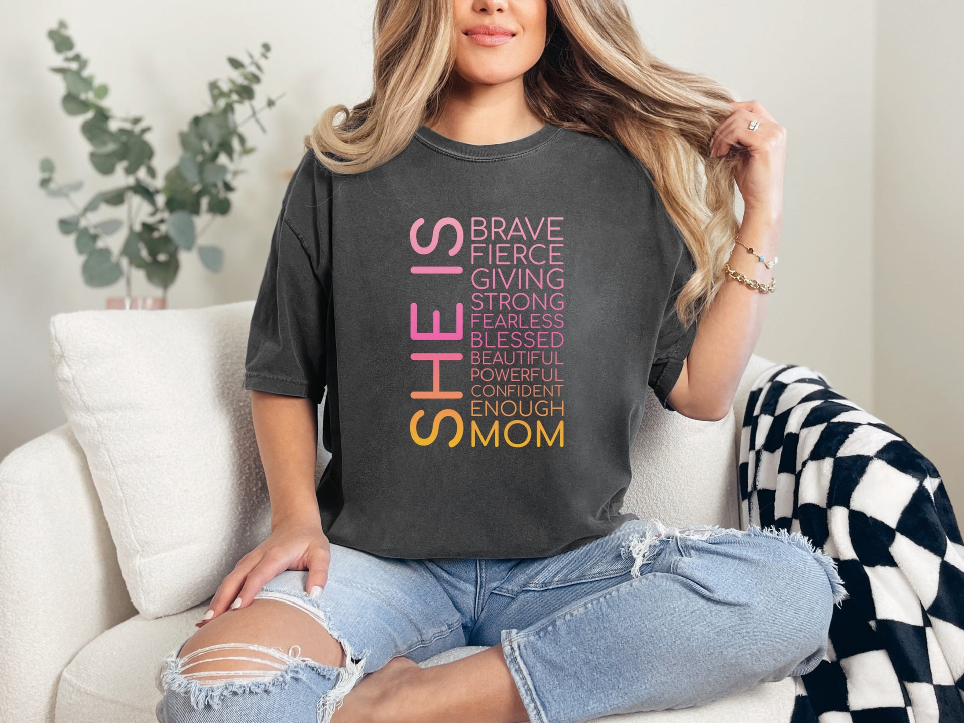 SHE IS vertical affirmation mom T-Shirt worn by a woman sitting on a couch, featuring uplifting affirmations.
