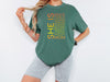Model wearing SHE IS vertical affirmation mom T-Shirt in green, showcasing uplifting words about motherhood.