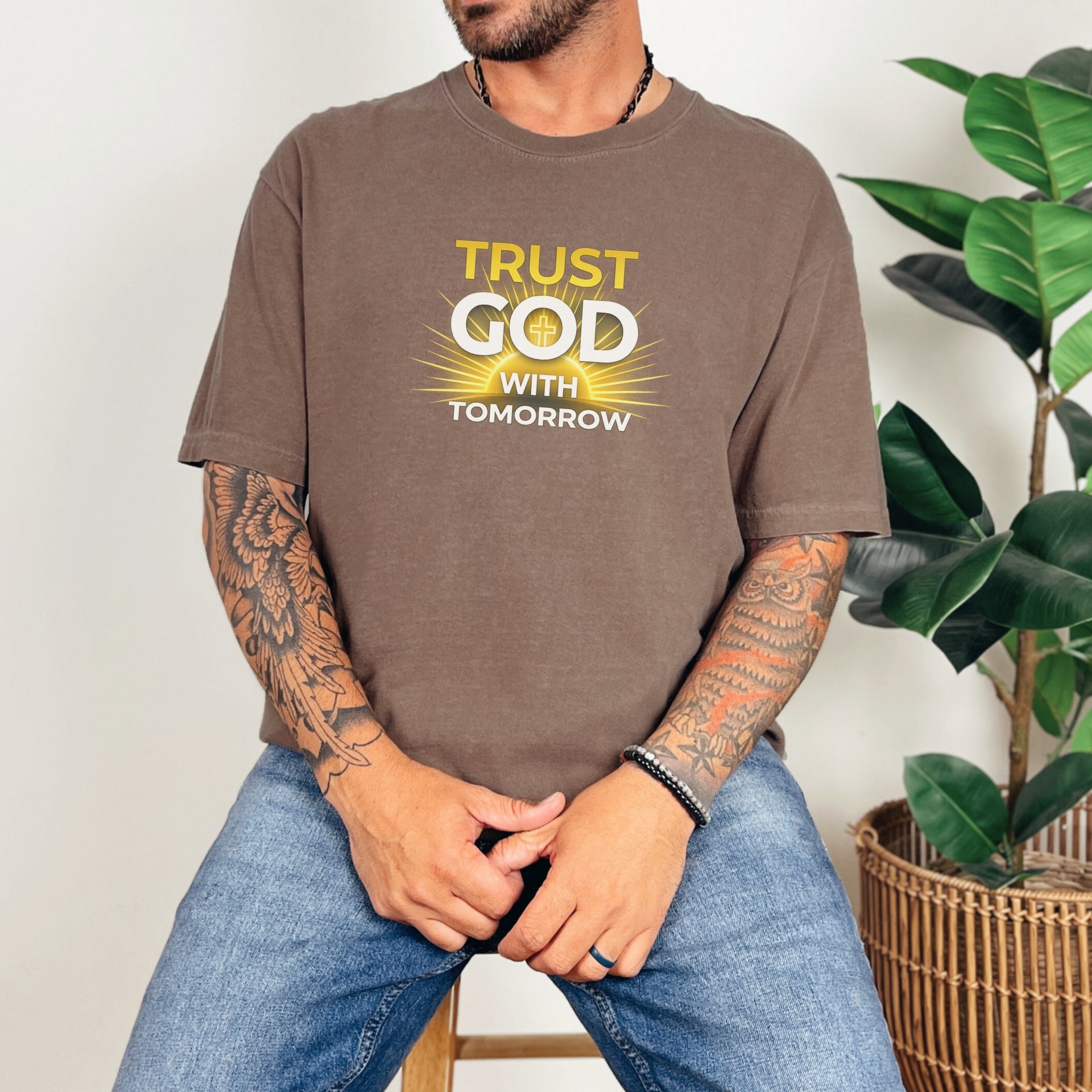 Model wearing Trust God With Tomorrow T-Shirt, featuring a motivational design in a casual setting with plants.