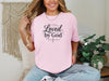 Woman wearing a pink Loved by God T-Shirt, showcasing faith and style in a cozy indoor setting.