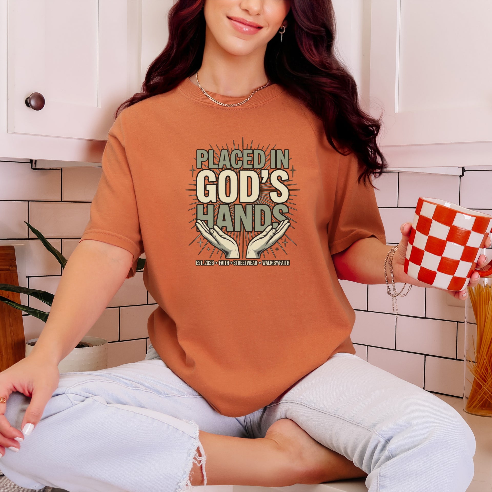 Woman wearing a Placed in God's Hands T-Shirt while holding a checkered cup in a cozy kitchen.