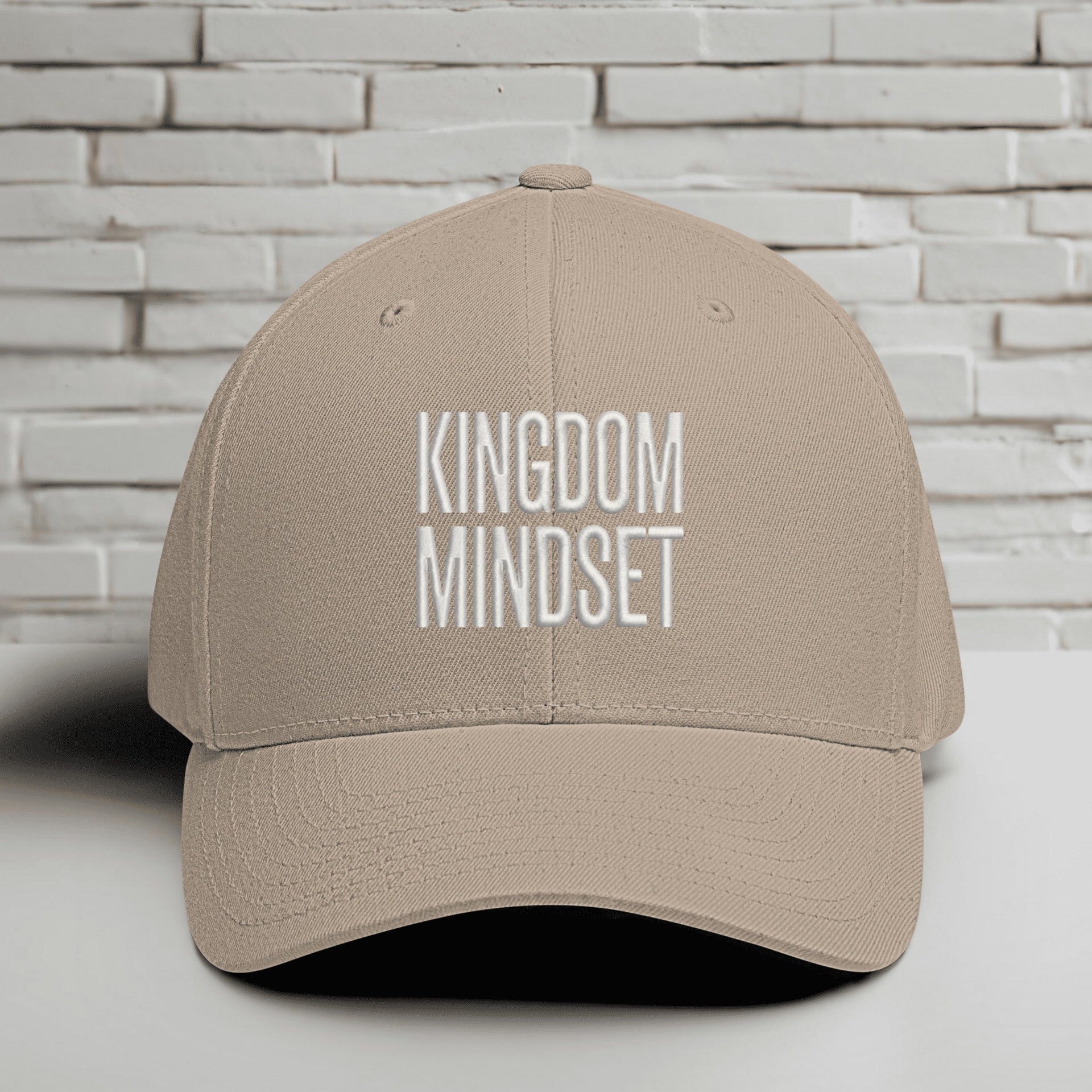 Kingdom Mindset Embroidered Flexfit Hat displayed on a flat surface against a brick wall background.