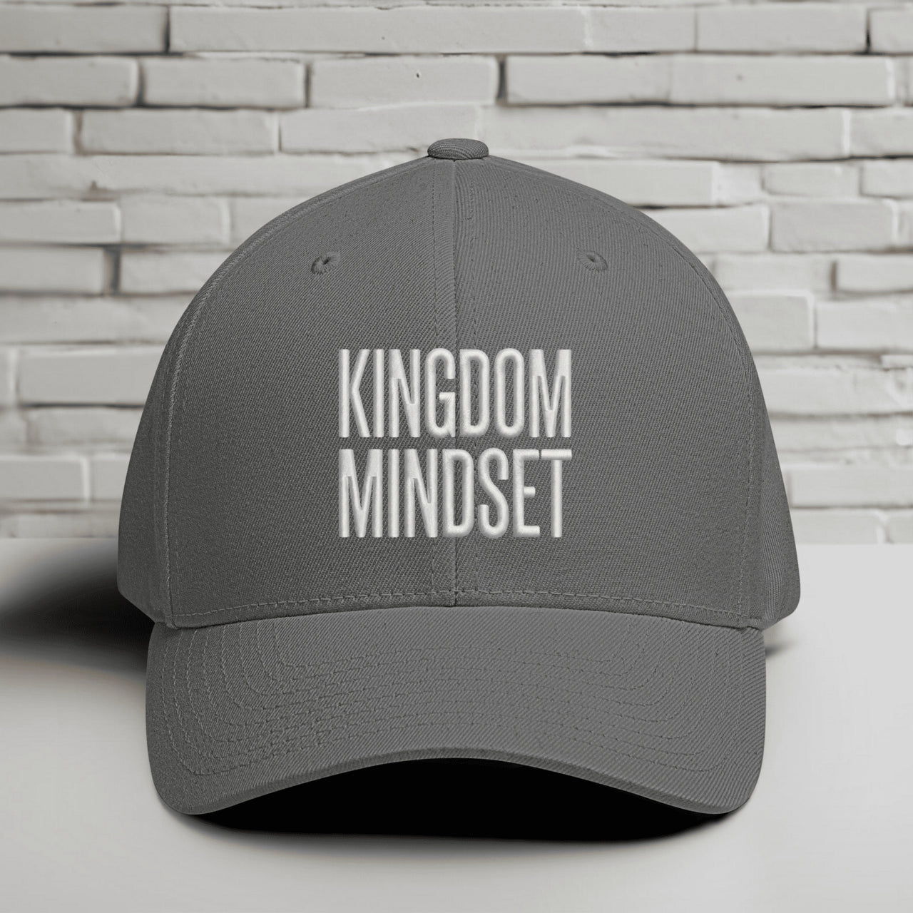 Kingdom Mindset Embroidered Flexfit Hat displayed on a table against a white brick background.