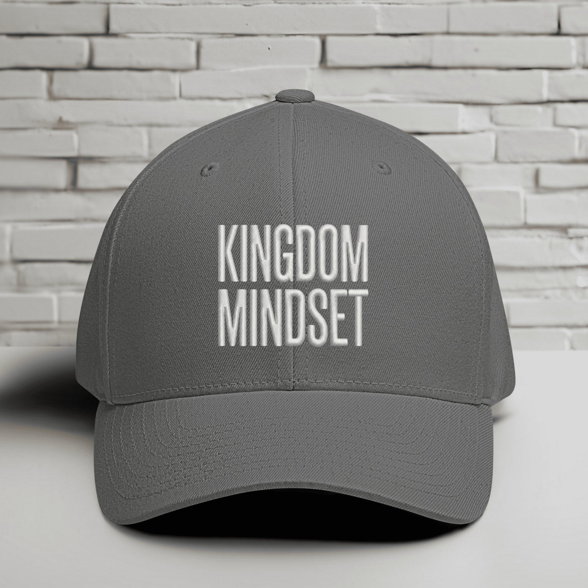Kingdom Mindset Embroidered Flexfit Hat displayed on a table against a white brick background.