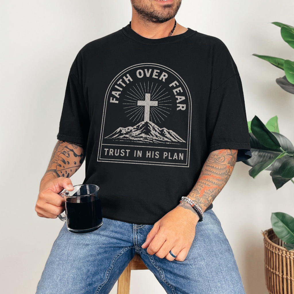 Man wearing a black Faith Over Fear Trust In His Plan Comfort Colors T Shirt, holding a cup, surrounded by plants.