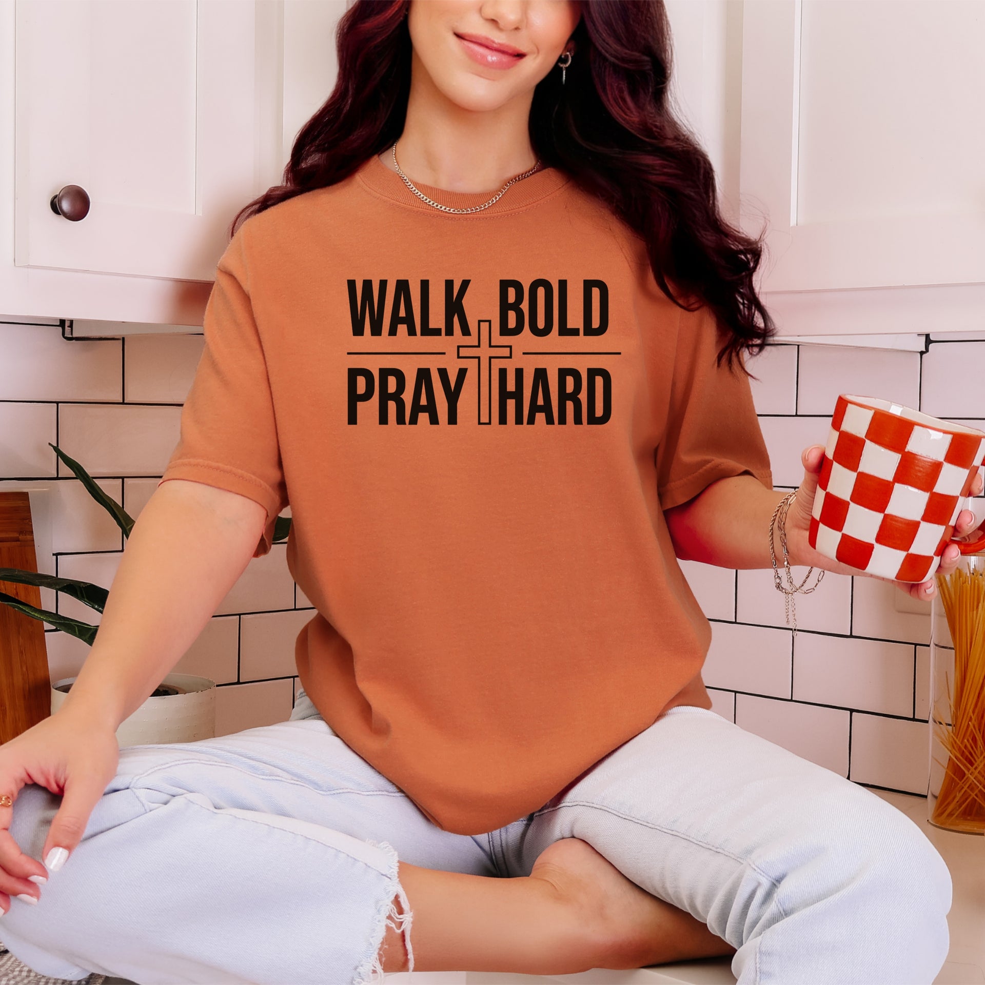 Woman wearing a Walk Bold Pray Hard Christian cross t shirt while sitting with a coffee mug in a kitchen.