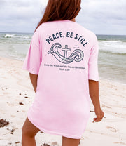 Woman wearing a pink Peace Be Still T Shirt at the beach with calming waves and a cross design.