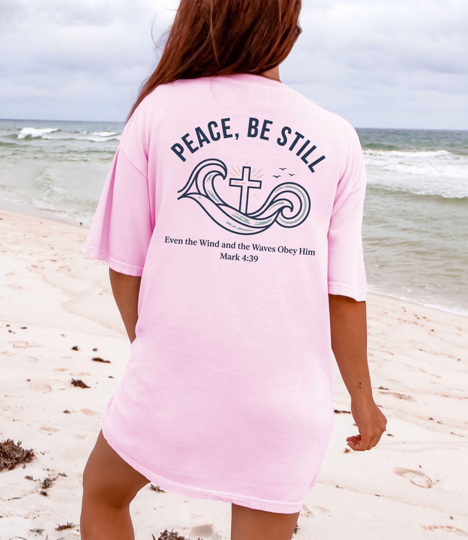 Woman wearing a pink Peace Be Still T Shirt at the beach with calming waves and a cross design.