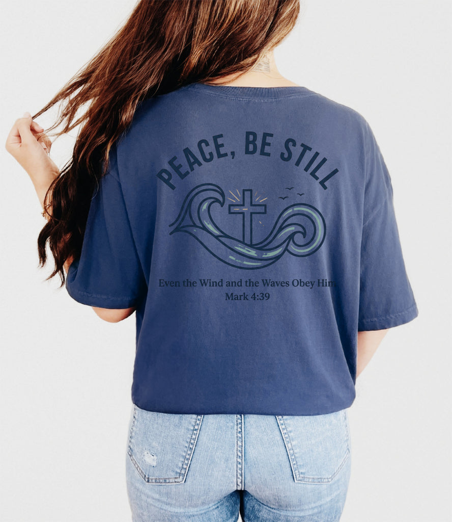 Woman wearing a navy Peace Be Still T Shirt with waves and cross design on the back, featuring Mark 4:39.