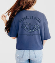 Woman wearing a navy Peace Be Still T Shirt with waves and cross design on the back, featuring Mark 4:39.