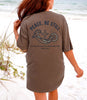 Peace Be Still T Shirt displayed on a woman at the beach, featuring a calming design with a cross and waves.