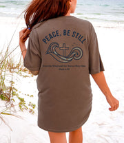 Peace Be Still T Shirt displayed on a woman at the beach, featuring a calming design with a cross and waves.