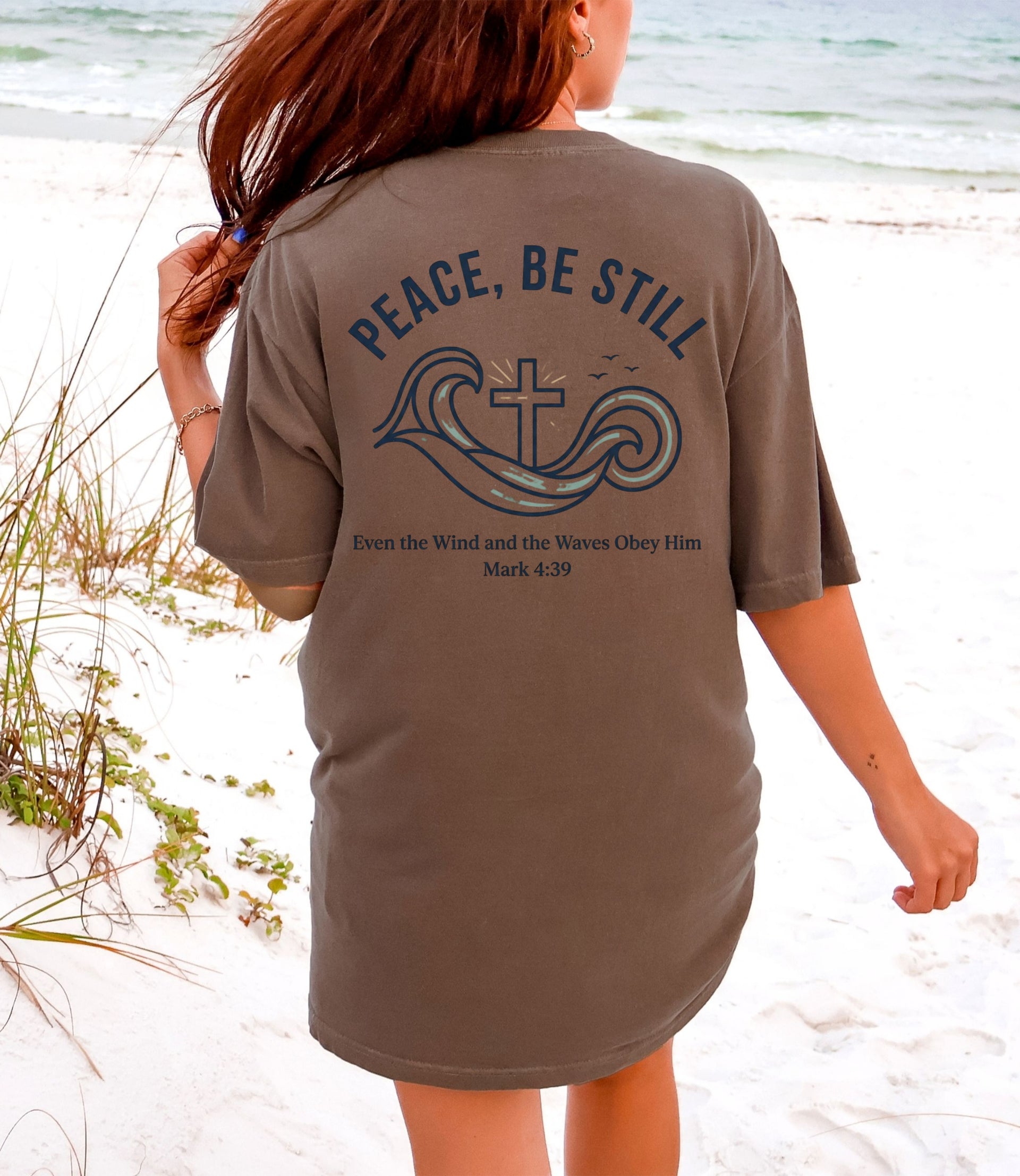 Peace Be Still T Shirt displayed on a woman at the beach, featuring a calming design with a cross and waves.