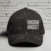 Flexfit Kingdom Mindset hat featuring clean white embroidery on a Multicam Black cap against a brick background.