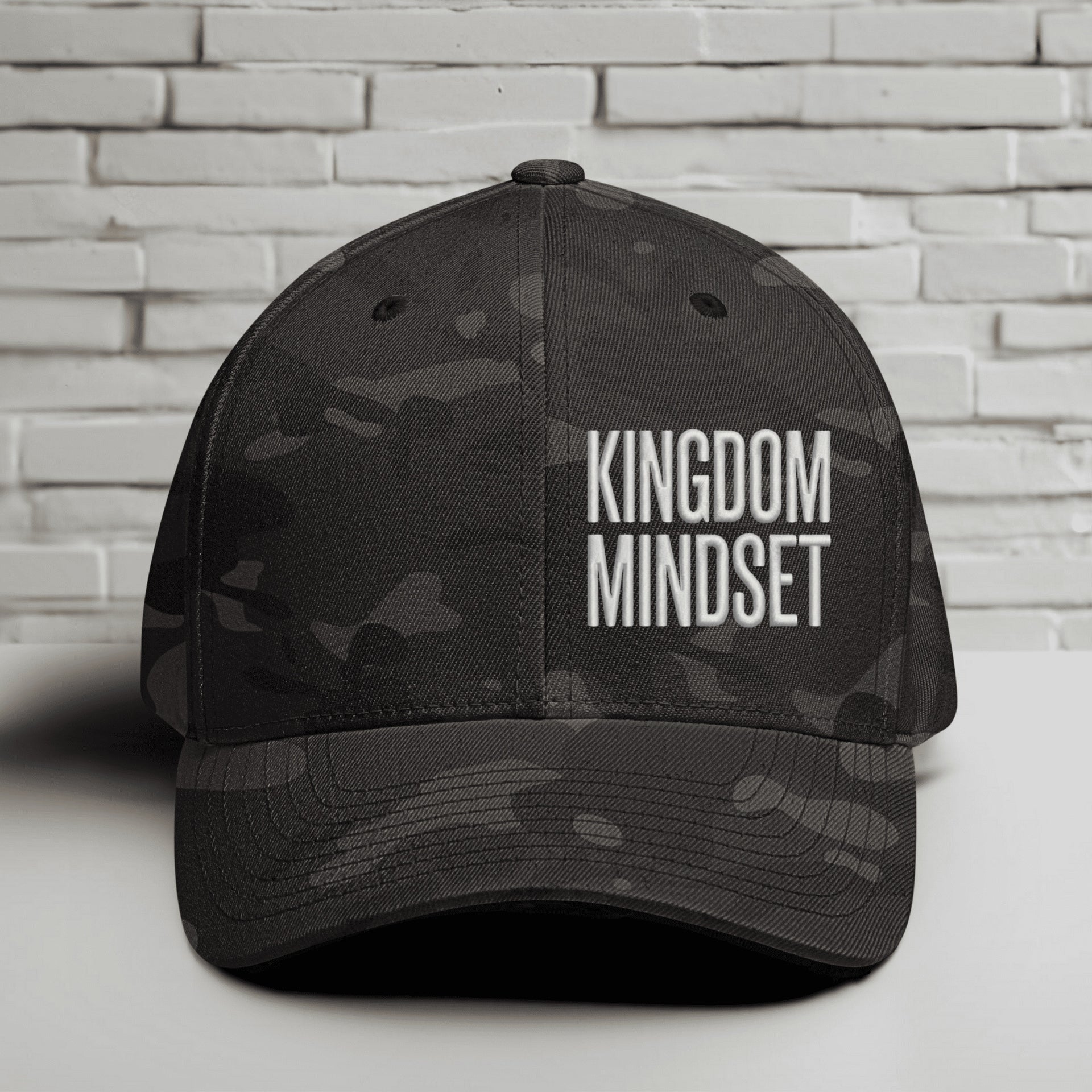 Flexfit Kingdom Mindset hat featuring clean white embroidery on a Multicam Black cap against a brick background.
