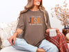 Woman wearing Fall for Jesus graphic tee in espresso color, cozy autumn setting with a coffee mug.