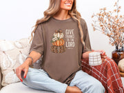 Woman wearing Give Thanks to the Lord t-shirt in espresso color, cozy autumn setting with pumpkins and coffee.