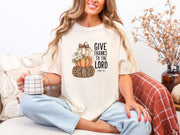 Woman wearing an ivory Give Thanks to the Lord T-Shirt with pumpkins, sipping coffee, celebrating gratitude and faith.
