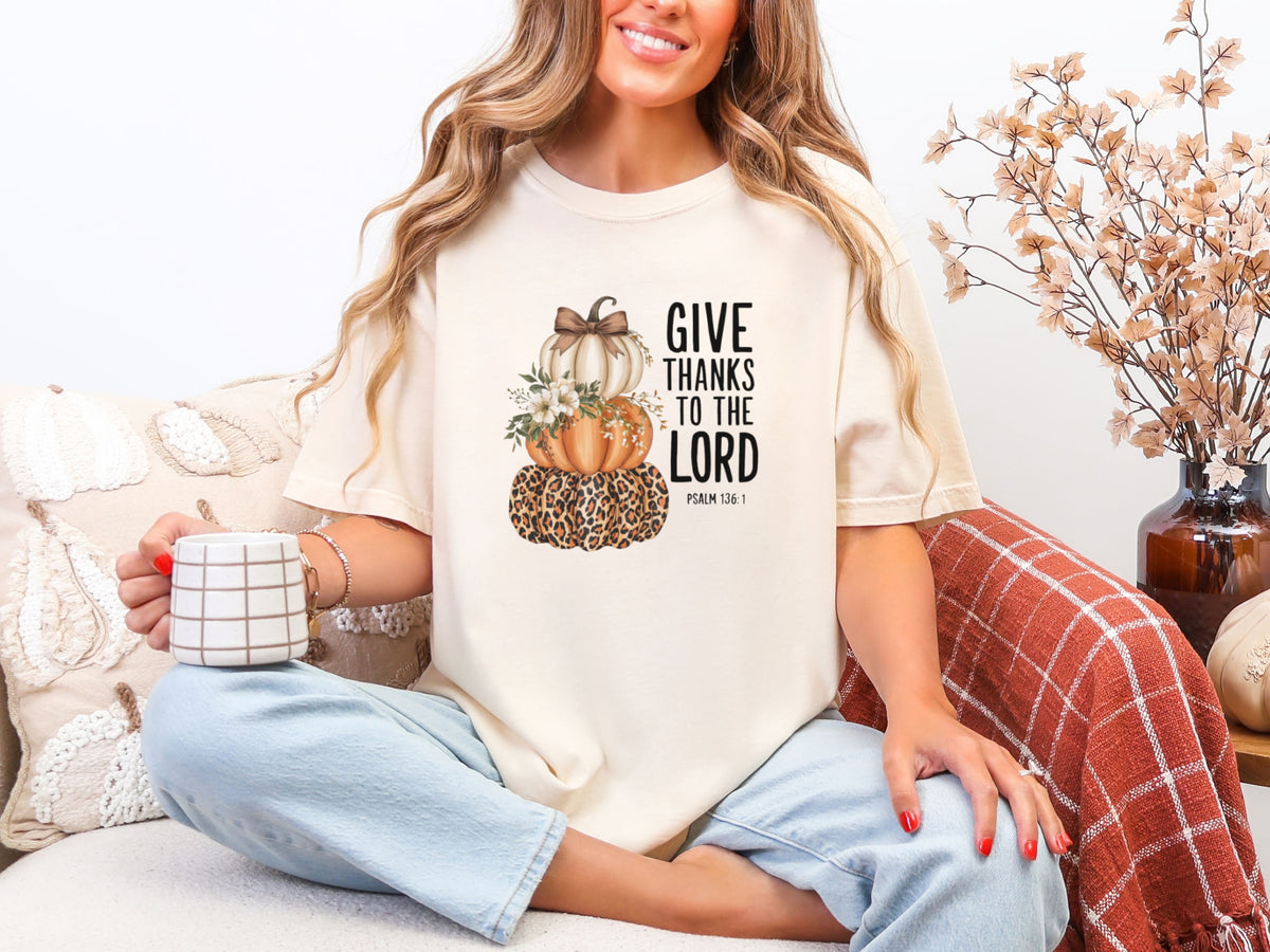 Woman wearing an ivory Give Thanks to the Lord T-Shirt with pumpkins, sipping coffee, celebrating gratitude and faith.