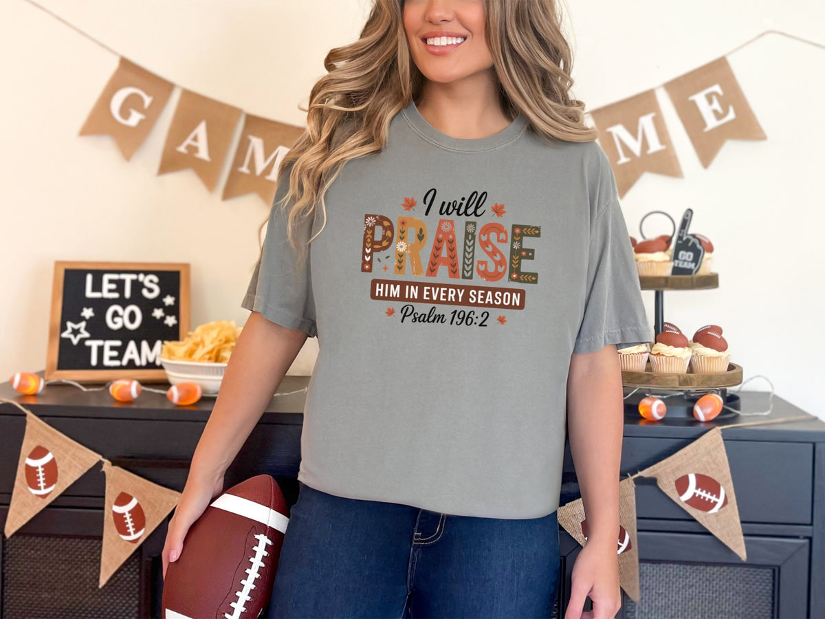 Woman wearing grey Christian graphic tee praise, featuring 'I Will Praise Him in Every Season' surrounded by autumn decor.