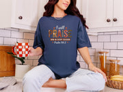 Woman wearing a navy I Will Praise Him in Every Season Christian graphic tee praise, holding a coffee mug in a cozy kitchen.