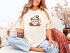 Woman enjoying coffee in a Jesus Fills My Cup t-shirt, paired with light blue jeans and seated on a cozy sofa.