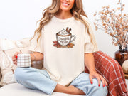 Woman enjoying coffee in a Jesus Fills My Cup t-shirt, paired with light blue jeans and seated on a cozy sofa.