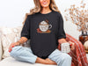 Woman wearing Jesus Fills My Cup t-shirt in black, holding a coffee mug, relaxing on a cozy sofa.