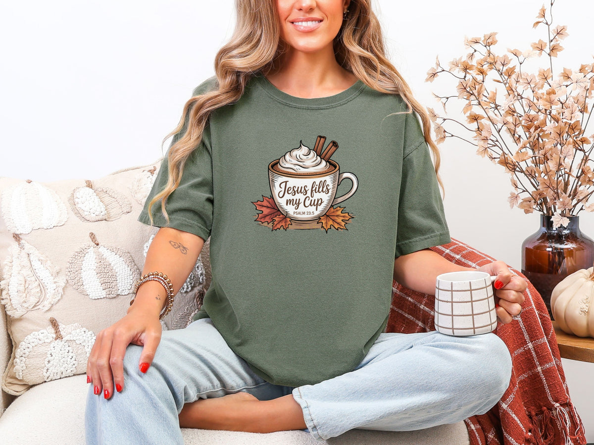 Woman wearing Jesus Fills My Cup t-shirt in moss color, holding a coffee cup, surrounded by cozy fall decor.