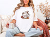 Woman wearing a white Jesus Fills My Cup t-shirt, sipping coffee, surrounded by cozy decor.