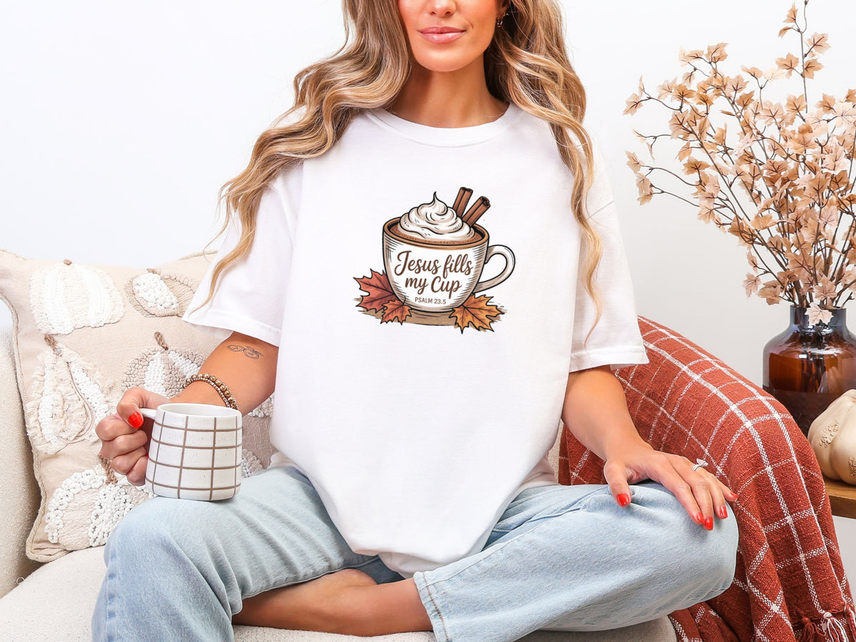 Woman wearing a white Jesus Fills My Cup t-shirt, sipping coffee, surrounded by cozy decor.
