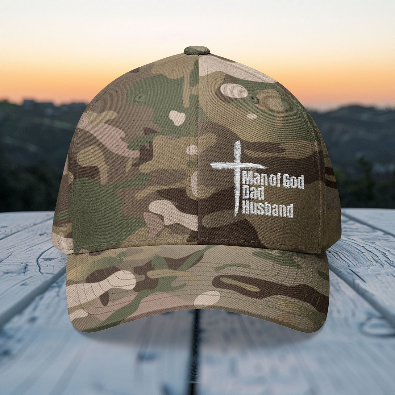 Man of God Dad Husband Embroidered Flexfit Hat in camouflage design with cross on outdoor wooden surface.