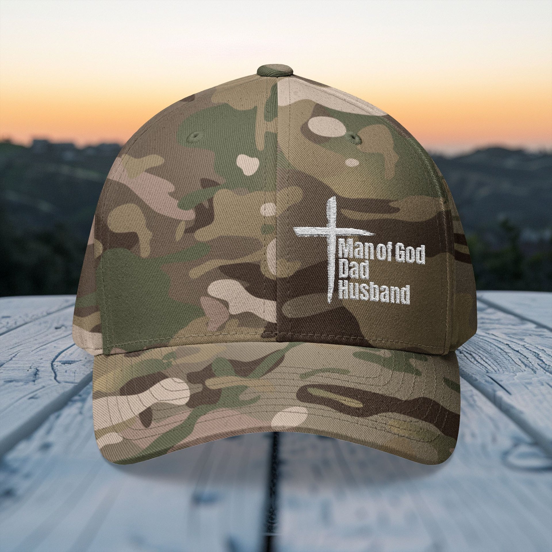 Man of God Dad Husband Embroidered Flexfit Hat in camouflage design with cross on outdoor wooden surface.